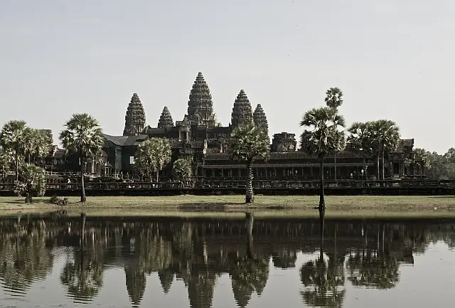 Antike Tempelanlage mit fünf pagodenartigen Türmen, eine Garten mit Palmen davor, an der Bildunterkante Wasser mit Spiegelung der Anlage