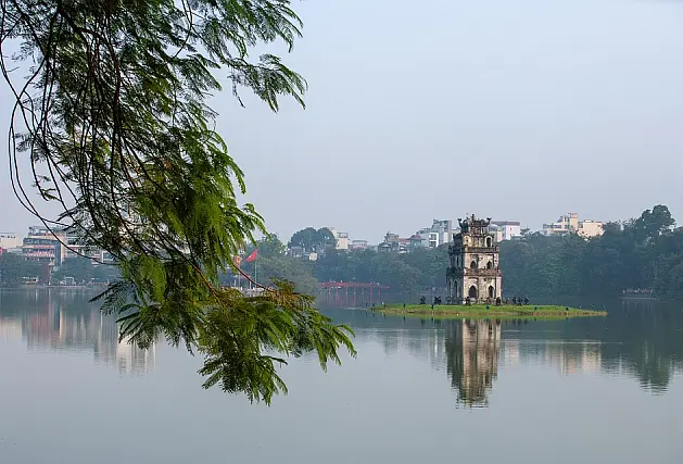 See mit kleiner Insel, auf der ein dreistöckiger Turm steht, im Hintergrund eine Stadt, im Vordergrund ein Zweig mit gefiederten Blättern