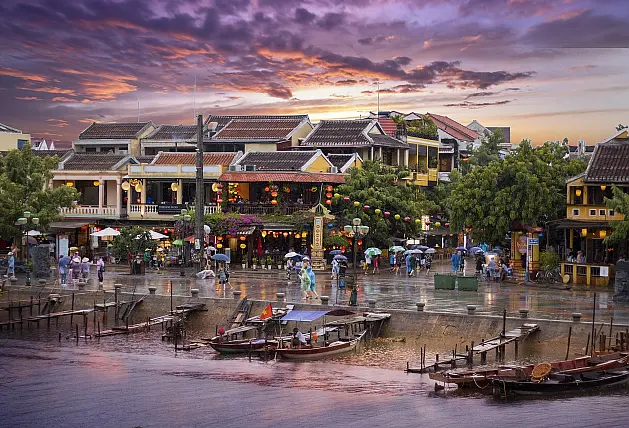 Blick vomMeer auf einen Fischerhafen mit kleinen Booten an den Stegen, belebte Straße und Fußgänger mit Regenschirmen im Regen, dahinter mehrstöckige Gebäude mit Lampions geschmückten Balkonen bei Sonnenuntergang mit violettem Himmel und orangefarbenen und violetten Wolken
