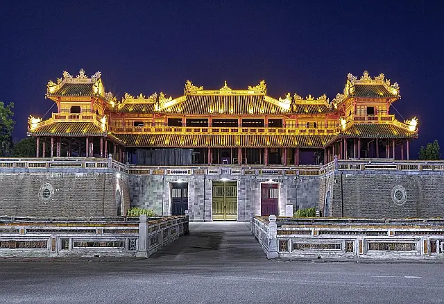 Altes vietnamesisches Steingebäude bei Nacht, mit beleuchtetem und kunstvoll verziertem Dach, im Vordergrund eine Brücke mit Steingeländer
