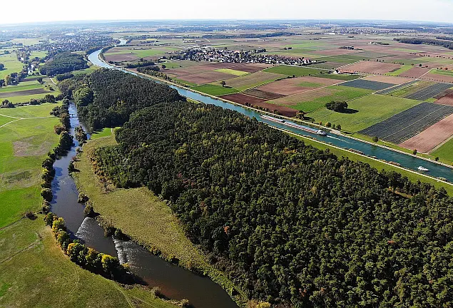 Luftbild mit Waldlandschaft, grünen und braunen Äckern, zwei Flüssen und einer Ortschaft im Hintergrund