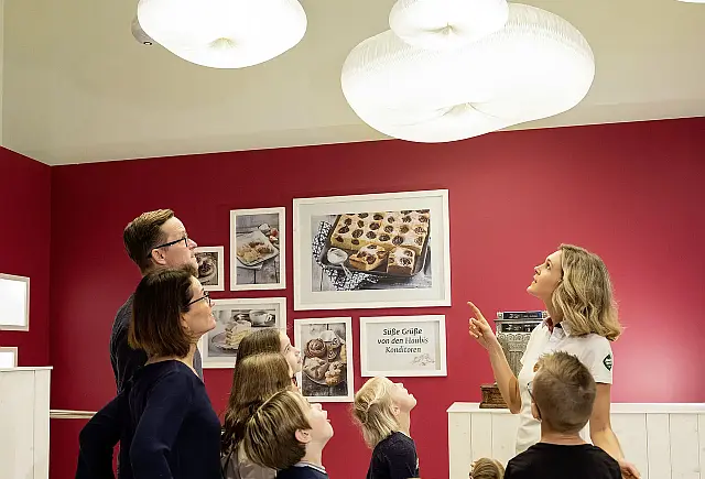 Familie mit Kindern unter weißen ovalen Deckenlampen stehend bei einer Gästeführung vor roter Wand mit eingerahmten Bildern von Kuchen und Mehlspeisen