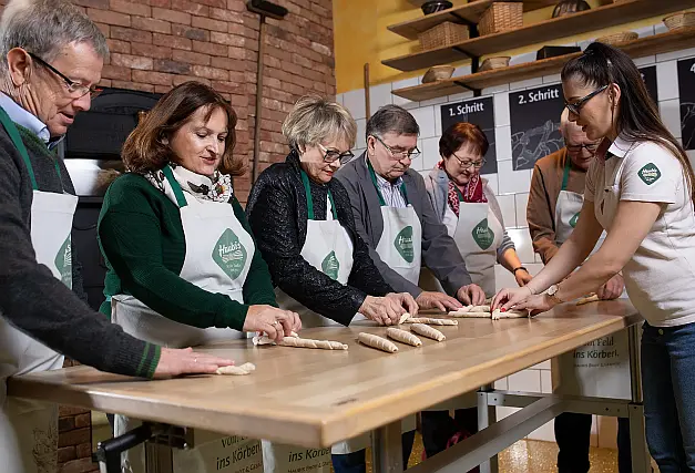 Männliche und weibliche Gäste mit weißen Kochschürzen und grünem Haubiversum-Logo rollen Teigstangen in einer Backstube mit roter Ziegelwand
