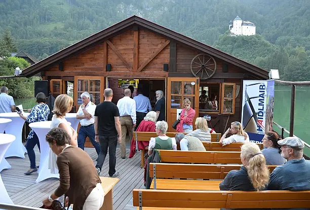Schwimmende Almhütte auf der Enns mit Gästen an Tischen und auf Bänken