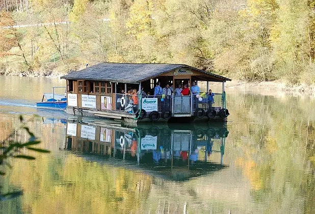 Schwimmende Almhütte fährt auf einem Fluss entlang, mit bewaldeter Uferböschung im Hintergrund