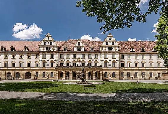 Ausschnitt einer Außenansicht eines Schloss mit historischer Fassade, rotem Dach mit Giebelfenstern und einem Park mit Springbrunnen im Vordergrund