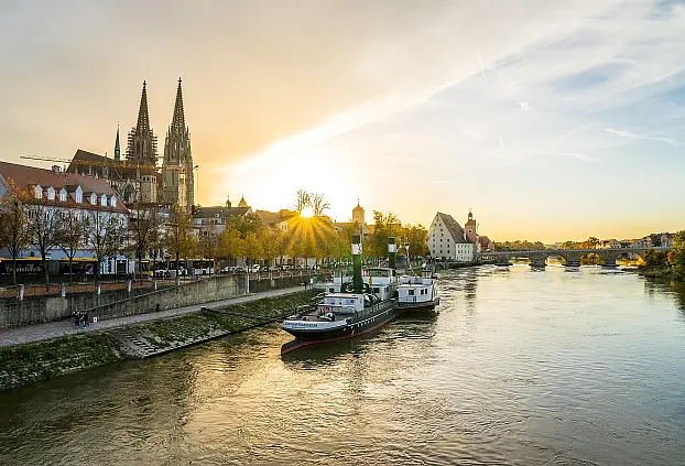 Fluss im Sonnenuntergang, ein Schliff liegt am Kai, ein Dom mit zwei Türmen und mehreren Gebäuden entlang des Kais, Brücke über Fluss am Horizont