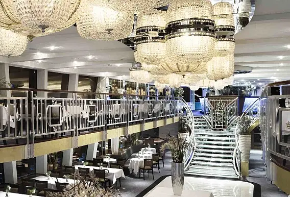 Speisesaal mit Kristallluster, Galagedeck und beleuchtetem Stiegenaufgang im Schiffsinnenbereich