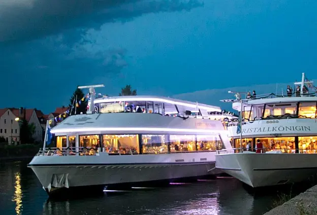 Zwei beleuchtete Schiffe bei Dämmerung, angelegt am Kai an einem Fluss, im Hintergrund zwei sind mehrstöckige Häuser zu erkennen