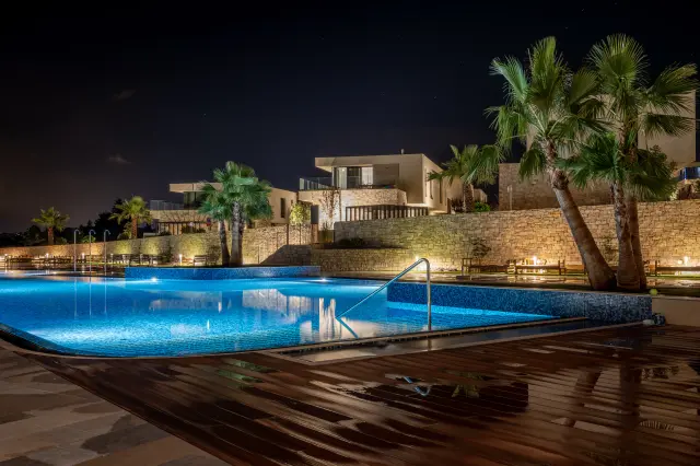 Beleuchteter Außenpool mit blau gefliestem Becken und nasser Holzterrasse bei Nacht im Bildvordergrund, dahinter entlang der Bildmitte eine Steinmauer mit beleuchteten Sitzbänken und Palmen, dahinterliegend zweistöckige Bungalows mit Flachdach, eingesäumt von Palmen