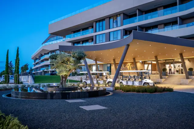 Überdachter Eingangsbereich mit Deckenbeleuchtung und zwei leeren Golf-Carts für Personenbeförderung vor einem Stiegenaufgang, mehrere tragende Säulen, dahinter mehrstöckiger Hotelkomplex, im Vordergrund ein Kiesweg und ein Brunnen mit beleuchtetem Baum in der Mitte, zwei Säulenzypressen am linken Bildrand