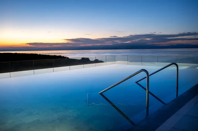 Infinity Pool bei Sonnenuntergang, mit blauem Wasser und Spiegelung des Himmels, Einstiegsgeländer an der rechten Bildecke, Blick auf das Meer am Horizont und einem Waldstück am linken Bildrand