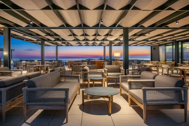 Gedämpft beleuchtete Hotellounge-Innenansicht mit länglichen beigefarbigen Bänden und kleine runden Tischen, eine geschlossener Dachbeschattung, mit Meerblick in der regenbogenfarbigen Abenddämmerung