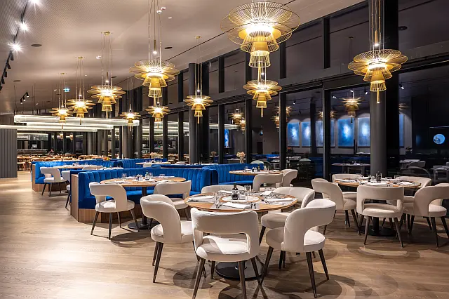 Innenansicht eines Speisesaales in einem luxuriös wirkendem Restaurant, leuchtende Kristallluster von der Decke hängend, gedeckte Tische mit runden weißen gepolsterten Stühlen, blau gepolsterte Sitzecken, Fensterfassade mit dunklen Rahmen entlang der rechten Saalseite