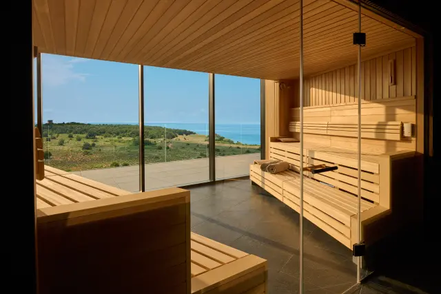 Sauna Innenbereich aus Holz, mit Glasfront im Vordergrund, mit Blick durch eine Glaswand auf das Meer und die Landschaft im Hintergrund