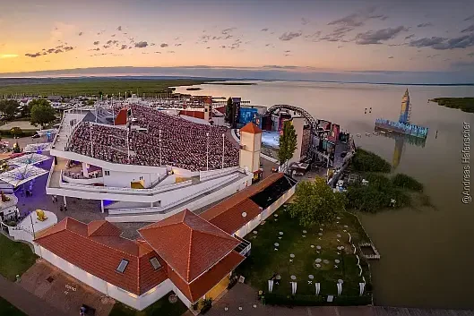 Drohnenfoto bei Sonnenuntergang, von einer Menschen befüllte ansteigende Tribüne an einem Gewässer liegend auf der rechten Bildseite, rote Dachflächen, grüne Wiesen und Bäume sowie beleuchtete Schirme am linken Bildrand, bei Sonnenuntergang