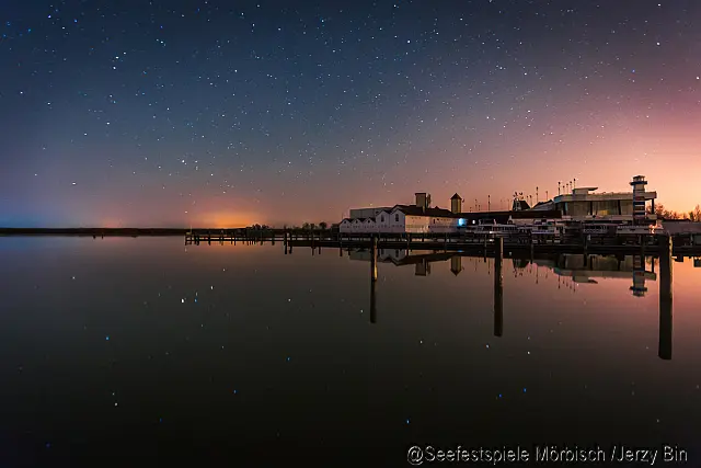 Blick vom Wasser auf mehrere niedrige Hafengebäude mit einem Leuchtturm an der rechten Bildkante, Holzstegen rundherum, bei Sonnenuntergang und Sternenhimmel