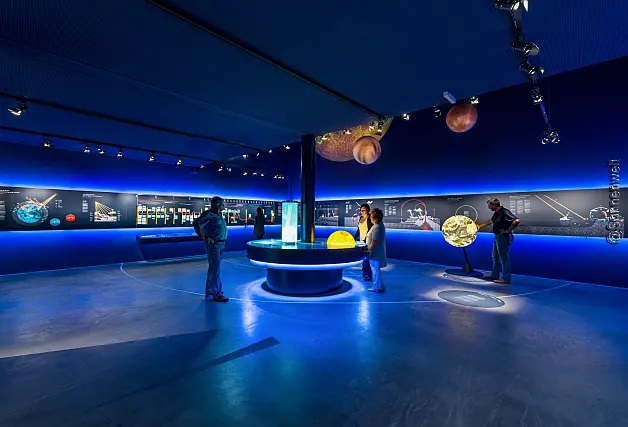 Ein dunkelblau beleuchteter Raum mit Besuchern, Wand-Screens und Konsolen mit interaktiven und multimedialen Ausstellungen über das Sonnensystem