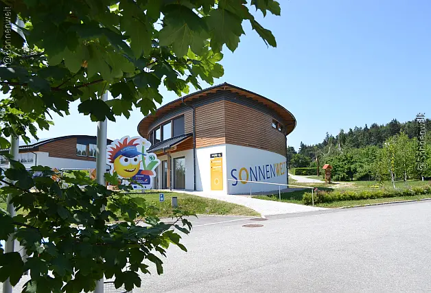 Zweistöckiges Gebäude in Form eines Digons mit Holzfassade am oberen Stockwerk und Sonnenwelt-Schriftzug, davorstehend ein Logo inmitten von Grünflächen mit Bäumen, Straße und Baumzweig mit grünen Blättern im Bildvordergrund