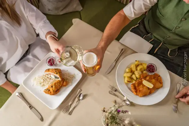 Bildausschnitt mit Blick von oben auf einen gedeckten Tisch mit einer weiblichen und männlichen Person, zwei Hände mit je einem Bierglas und einem Weinglas, welche die Gläser zusammenstoßen und zwei mit Speisen gefüllte Teller