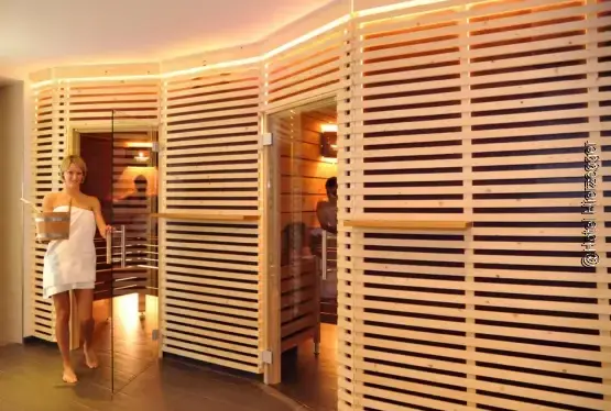 Außenansicht von zwei Saunakabinen aus Holz mit Glastüren, gelber indirekter Beleuchtung und einer Dame in Badetuch gewickelt mit Aufgussfass stehend vor der linken Saunatür