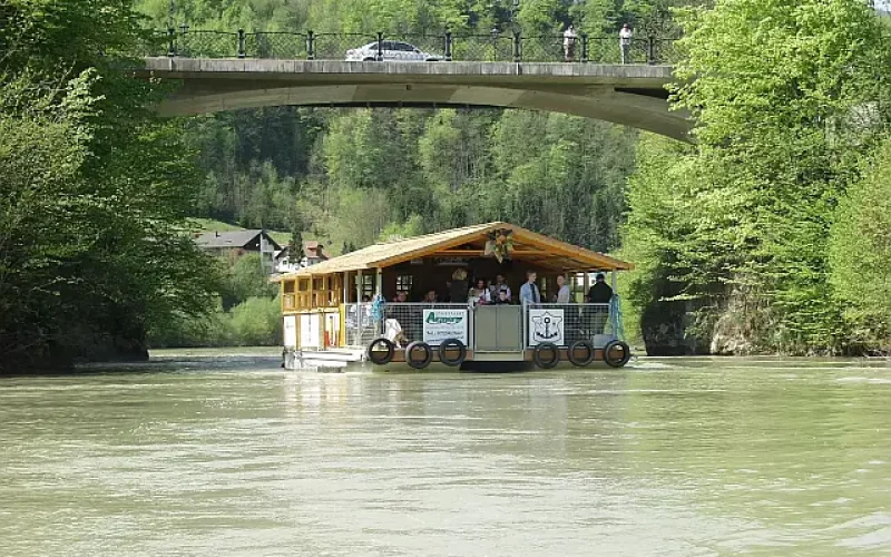 Schwimmendes Boot mit Almhütte auf einem bewaldeten Flusstal durchquert eine Brücke, auf der ein PKW und 2 Personen am Brückengelände stehen